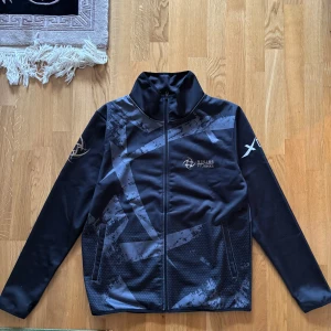 Svart jacka från Ninjas in Pyjamas - Säljer en svart jacka från Ninjas in Pyjamas med coolt mönster och tryck på både fram- och baksidan. Jackan har dragkedja och långa ärmar, perfekt för dig som gillar gaming och stil. #GONINJAS