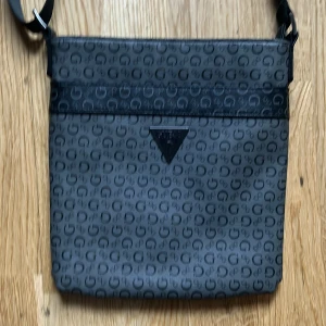 Svart axelväska från Guess - Snygg svart axelväska från Guess med ett stilrent mönster av små G:n. Väskan har en justerbar axelrem och en diskret Guess-logga i metall på framsidan. Perfekt för att bära dina dagliga nödvändigheter med stil.