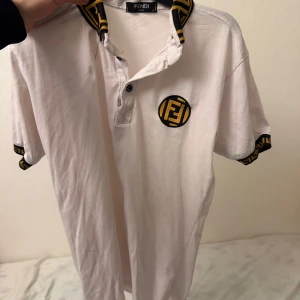 Vit kortärmad t-shirt från Fendi - Säljer en stilren vit t-shirt från Fendi med korta ärmar. T-shirten har en svart och gul mönstrad krage och ärmslut med Fendi-logga. Perfekt för en trendig look.