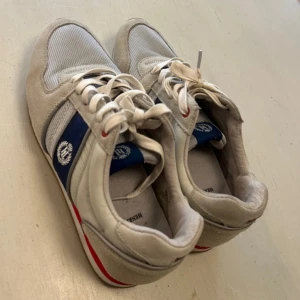 Vita/ljusgråa sneakers från Henri Lloyd stl 44 - Snygga vita sneakers från Henri Lloyd med blå och röda detaljer. Skorna har snörning och en kombination av mocka och syntetmaterial. Perfekta för en stilren look.storlek 44