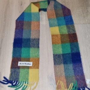 Rutig halsduk från Acne Studios - Snygg och färgglad rutig halsduk från Acne Studios. Den har ett mönster i blått, grönt, gult och orange med fransar i ändarna. Perfekt för att liva upp din outfit!