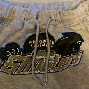 Grå mjukisbyxor från Trapstar - Snygga grå mjukisbyxor från Trapstar med ett coolt tryck på framsidan. Byxorna har snörning i midjan för justerbar passform och är perfekta för en avslappnad stil. De är tillverkade i ett mjukt och bekvämt material.