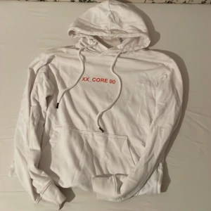 Vit hoodie från Jack & Jones med tryck - Säljer en vit hoodie med texten 'XX_CORE 90' på framsidan och ett stort tryck med stadsmotiv på baksidan. Hoodien har en klassisk känguruficka och justerbar huva med dragsko. Perfekt för en avslappnad stil.