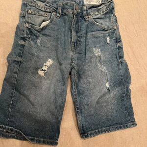 Blå jeansshorts från 157 - Snygga blå jeansshorts från 157 med slitna detaljer för en cool look. De har en klassisk femficksdesign och knappgylf. Perfekta för en avslappnad stil. (St 150)