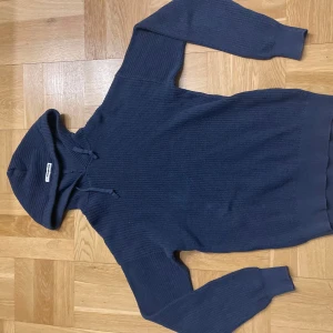 Mörkblå hoodie från REBEL - Säljer en snygg mörkblå hoodie från REBEL i storlek S. Den är stickad med en ribbad struktur och har en klassisk huva med dragsko. Perfekt för kyliga dagar!