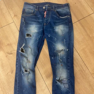 Blå jeans med slitningar från Dsquared2 äkta - Snygga blå jeans från Dsquared2 äkta med coola slitningar och färgstänk. De har en klassisk femficksdesign och en röd logga på bakfickan. Perfekta för en avslappnad stil med en edgy touch.