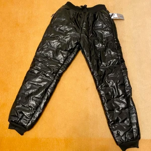 Ny Svarta vadderade coola täckbyxor - Snygga svarta täckbyxor med glansig yta och dragkedjor. Perfekta för kalla dagar med sin quiltade design. Byxorna har elastisk midja och muddar vid bensluten för extra komfort. Någon mindre nyansskillnad finns i material, men inget som syns. Storlek M eller lite större S.