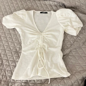 Vit blus från BikBok - Säljer en söt vit blus från BikBok med puffiga kortärmar och en v-ringad design. Blusen har en snygg snörning framtill som ger en fin detalj. Perfekt för att piffa upp garderoben!