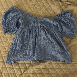 Blå blus med puffärmar - Charmig blå blus med puffiga kortärmar och smockdetaljer. Blusen har en snörning i ryggen som ger en fin passform. Perfekt för en somrig look!