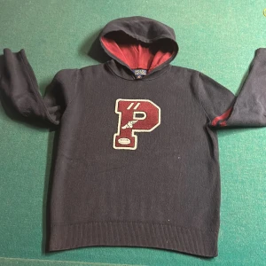 Svart hoodie från Polo Ralph Lauren - Säljer en svart hoodie från Polo Ralph Lauren med en stor röd bokstav 'P' på framsidan. Hoodien har en röd insida i luvan och ribbade muddar. Perfekt för en avslappnad stil.