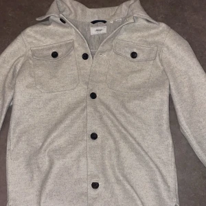 Grå overshirt från Produkt - Säljer en stilren grå overshirt från Produkt. Den har långa ärmar och knappar framtill samt två bröstfickor med knappar. Perfekt för lager-på-lager-stil. Stor i storleken skulle passa någon som är M-L inte S
