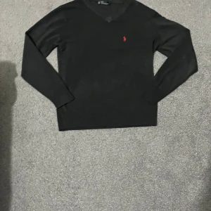 Svart kashmirtröja från Ralph Lauren - Säljer en elegant svart kashmirtröja från Ralph Lauren med en klassisk v-ringning. Tröjan har ett diskret rött broderat märke på bröstet och långa ärmar. Perfekt för en stilren look. Storlek L men passar M