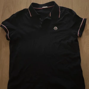 Svart pikétröja från Moncler - Snygg svart pikétröja från Moncler med klassisk krage. Tröjan har röd och vit randdetalj på krage och ärmslut samt Moncler-logga på bröstet. Priset är inte hugget i sten. Skriv om ni har funderingar.