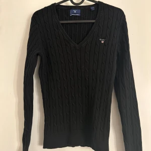 Svart ribbad tröja från GANT - Säljer en stilren svart ribbad tröja från GANT i stretchig bomull. Tröjan har en v-ringad hals och långa ärmar, perfekt för en klassisk look. Den är både bekväm och snygg, idealisk för kyligare dagar.