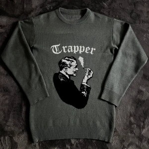 Made by trappers stickad  - Snygg grön tröja med ett unikt motiv av en man och texten 'Trapper' i vitt. Tröjan har långa ärmar och ribbade muddar vid ärmslut och nederkant. Bilden gör så att ena armen ser mörkare ut men den har exakt samma färg hela tröjan. Storlek L, pris går att diskutera✅