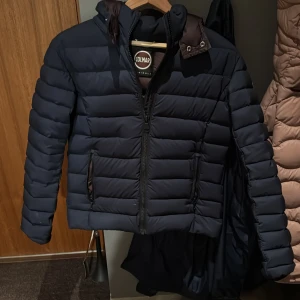 Mörkblå pufferjacka från Colmar - Säljer en snygg mörkblå pufferjacka från Colmar. Jackan har en dragkedja framtill och en praktisk huva. Perfekt för kyliga dagar med sin quiltade design som håller värmen. Den har långa ärmar och en stilren look. Tillkommer tags, storleken är xs-xxs och den passar en bra vid längden 165-170 bra skulle jag säga