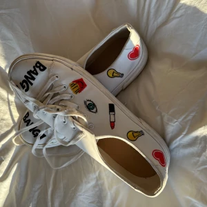 Vita sneakers med tryck - Unika vita sneakers med färgglada tryck av ögon, läppstift, hjärtan och texten 'BANG!'. Skorna har snörning och en klassisk design som sticker ut med sina lekfulla detaljer. Stl 38-40!