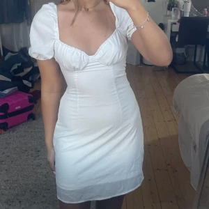 Vit kortklänning med puffärmar - Säljer en söt vit kortklänning med puffärmar och en smickrande v-ringad design. Klänningen har en tight passform och är perfekt för sommarens alla tillfällen. Den är gjord i ett lätt och bekvämt material som ger en fin siluett.