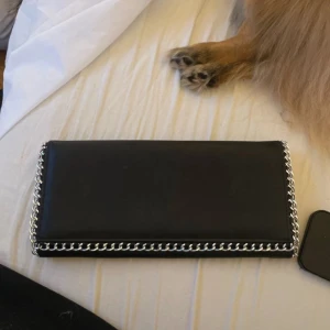 Svart kuvertväska från Gina Tricot - Snygg svart kuvertväska från Gina Tricot med kedjedetaljer längs kanterna. Väskan har en praktisk dragkedja inuti och stängs med en tryckknapp. Perfekt för att bära det viktigaste med stil.