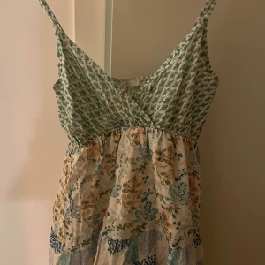 Blommig sommarklänning - Charmig klänning med grönt och blommigt mönster. Den har en omlott-design upptill och smala axelband. Perfekt för varma dagar med sitt luftiga material och vackra blommönster i olika färger.