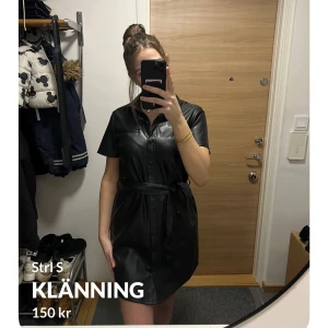 Svart klänning från H&M - Snygg svart klänning i syntetmaterial från Divided. Den har korta ärmar och ett bälte i midjan för en stilren look. Perfekt för en cool och edgy stil. Klänningen har knappar framtill och en klassisk krage.