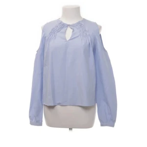 Ljusblå blus med öppna axlar - Säljer en ljusblå blus med öppna axlar och lång ärm. Blusen har en dekorativ öppning vid halsen och rynkade detaljer. Perfekt för en stilren look.
