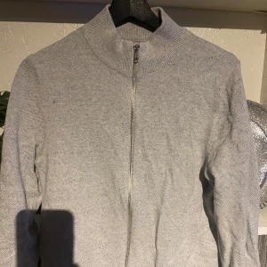 Grå tröja med dragkedja från Jack & Jones - Säljer en stilren grå tröja från Jack & Jones med dragkedja framtill. Tröjan har en hög krage och långa ärmar, perfekt för kyligare dagar. Passar bra till både jeans och chinos.