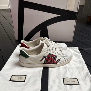 Vita sneakers från Gucci med ormmotiv - Snygga vita sneakers från Gucci med ett coolt ormmotiv på sidan. Skorna har klassiska gröna och röda ränder samt snörning. Tillverkade i Italien med högkvalitativt skinn. Väldigt bra skick sparsamt använda dustbag tillkommer vid eventuell överenskommelse måste ske en förfrågan alltså. Priset går att diskutera till en viss gräns. Mer frågor så hör gärna av dig mvh Axel.