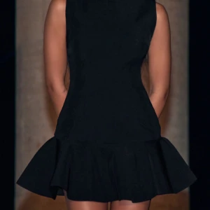 Svart kort klänning med volang - Snygg svart kort klänning med volangdetalj i nederkant. Klänningen är ärmlös och har en figurnära passform som framhäver silhuetten. Perfekt för en kväll ute eller en speciell tillställning. Aldrig använd! 