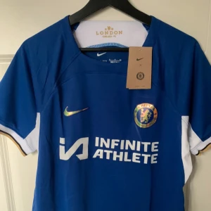 Blå Chelsea FC fotbollströja från Nike - Säljer en blå Chelsea FC fotbollströja från Nike med korta ärmar. Tröjan har klubbens emblem på bröstet och texten 'Infinite Athlete' tryckt på framsidan. Perfekt för alla Chelsea-fans! 💙