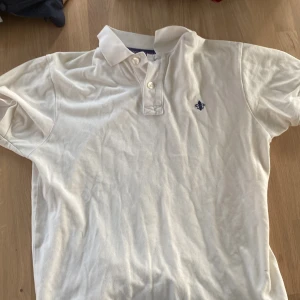 Vit pikétröja från Morris - Säljer en stilren vit pikétröja från Morris med en broderad blå detalj på bröstet. Tröjan har en klassisk krage och knappar framtill. Perfekt för en avslappnad men ändå snygg look.