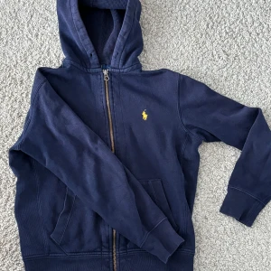 Mörkblå huvtröja från Polo Ralph Lauren - Säljer en snygg mörkblå huvtröja från Polo Ralph Lauren med dragkedja och det klassiska gula logotypbroderiet på bröstet. Tröjan har långa ärmar och en praktisk huva, perfekt för kyliga dagar. Den är tillverkad i ett mjukt och bekvämt material.