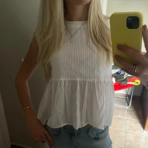 Vit topp - Säljer en söt vit blus med tunna ränder och volangdetalj nedtill. Den är ärmlös och har en luftig passform, perfekt för varma dagar. Passar bra till jeans eller kjol för en avslappnad look. XS men sitter helt som en S så passformen är lite lös