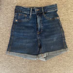 Säljer ett par snygga mörkblå jeansshorts med hög midja. De har en klassisk femficksdesign och är uppvikta vid bensluten för en trendig look. Perfekta för sommardagar eller en avslappnad utflykt. Passar till allt från t-shirts till blusar!