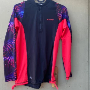 Svart och röd rashguard från Olaian - Säljer en snygg och funktionell rashguard från Olaian i svart och rött med coola mönstrade ärmar i lila och blått. Perfekt för soliga dagar med UPF 50+ skydd. Den har en dragkedja framtill och långa ärmar. Perfekt för surfing eller andra vattensporter!