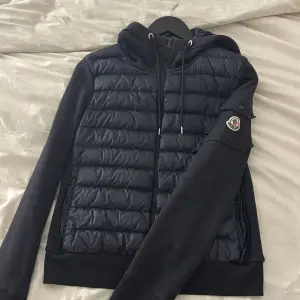 Säljer en snygg svart cardigan från Moncler. Den har en quiltad design med dragkedja och huva. Jackan är perfekt för höst och vinter, och den har långa ärmar. Den är i nyskick och superbekväm att ha på sig. Passar perfekt för en casual look 