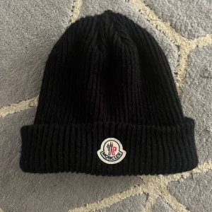Svart ribbad mössa från Moncler - Säljer en snygg svart ribbad mössa från Moncler. Den är perfekt för kalla dagar och har en klassisk stil med Monclers logga framtill. Mössan är både varm och stilren, vilket gör den till ett måste i vintergarderoben. 10/10 skick 