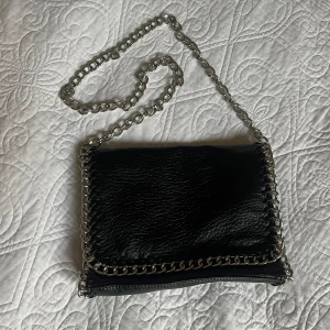 Svart axelväska med kedjedetaljer - Säljer en svart väska från Tiamo i konstskinn med coola kedjedetaljer. Endast använd ett fåtal gånger. Väskan har ett fack med dragkedja på baksidan och flera mindre fack/innerfickor. Mått ca: B=28 cm, H=20 cm, D=5,5 cm. Axelkedjans längd ca 109 cm