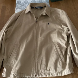 Beige jacka från Polo Ralph Lauren - Säljer en snygg beige jacka från Polo Ralph Lauren. Den har en klassisk design med dragkedja och två fickor framtill. Jackan är perfekt för vår och höst, och den har en stilren look med det ikoniska Polo Ralph Lauren-märket på bröstet.