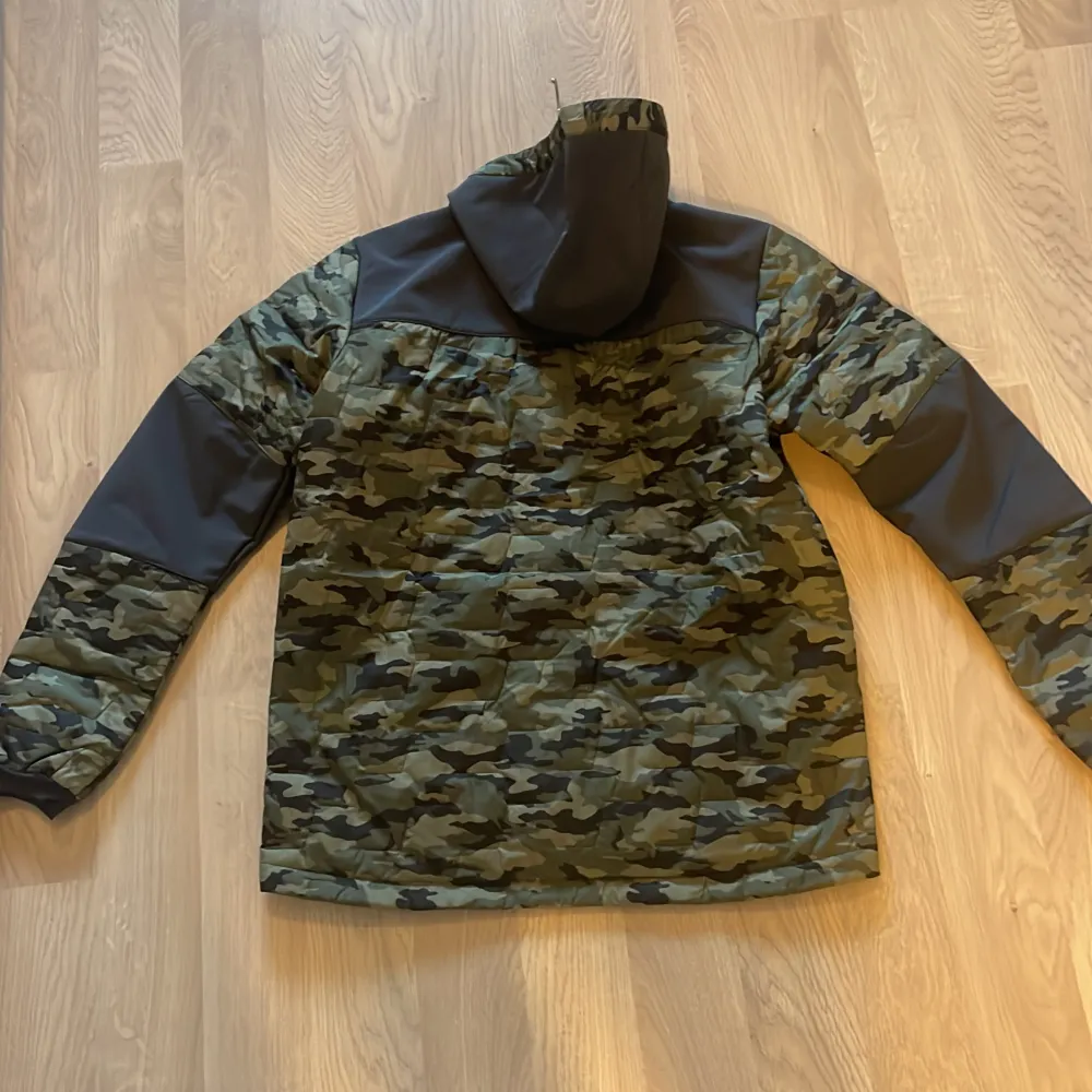 Säljer en snygg och praktisk camouflagejacka från Helly Hansen. Jackan har en cool camo-design med svarta detaljer på ärmarna och axlarna. Den är långärmad och har en dragkedja framtill. Perfekt för höst och vår, och den är i nyskick. Passar bra för utomhusaktiviteter och har en normal passform.. Jackor.