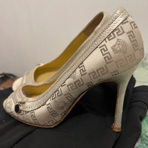 Beige pumps från Christian Louboutin - Säljer ett par eleganta beige pumps från Christian Louboutin. Skorna har en spetsig tå och en hög klack, perfekt för festliga tillfällen. De är dekorerade med ett subtilt mönster och har en liten metallisk detalj framtill. Materialet ser ut att vara en blandning av skinn och syntet, vilket ger dem en lyxig känsla.