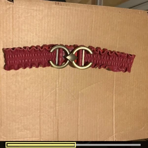 Rött bälte från Saddler - Säljer ett snyggt rött bälte från Saddler i äkta läder. Bältet har en cool design med ett elastiskt, rynkat band och ett stort guldfärgat spänne i form av två sammanlänkade C:n. Perfekt för att ge din outfit en extra touch! Passar midjemått på 75 cm.