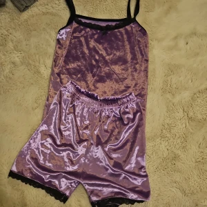 Lila sammet pyjamasset - Säljer ett supermysigt lila pyjamasset i sammet. Setet består av ett linne med svarta spetsdetaljer och matchande shorts med elastisk midja. Perfekt för en avslappnad kväll hemma eller som en söt present! 💜