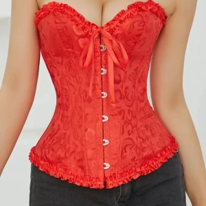 Röd korsett med snörning och volangdetaljer - Säljer en superfin röd korsett med snygg snörning framtill och volangdetaljer längs kanterna. Den har ett vackert mönster och knappar framtill så man kan få på den lättare🫶🏻 den passar dem flesta tack vare att man snarare den bak
