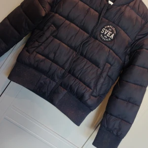 Marinblå pufferjacka från Svea - Säljer en svart pufferjacka från Svea i storlek 140. Jackan har en dragkedja framtill och är långärmad. Perfekt för höst och vinter med sin varma och bekväma design. Svea-loggan är snyggt placerad på bröstet.