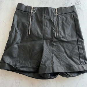 Svarta shorts med dragkedjor - Snygga svarta shorts i skinnimitation med coola dragkedjor framtill. Perfekta för en utekväll eller fest! De har en hög midja och en stilren design som passar till det mesta. Ett måste i garderoben för den som gillar att sticka ut.