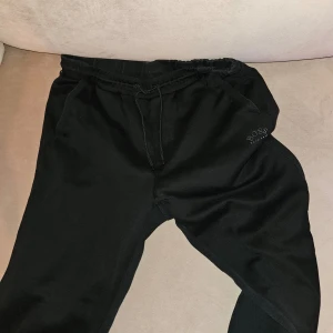 Svarta mjukisbyxor från Hugo Boss - Säljer ett par svarta mjukisbyxor från Hugo Boss. De har en bekväm passform med elastisk midja och snörning. Byxorna har fickor på sidorna och en diskret logga på ena benet. Nyskick.