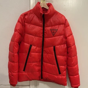 Röd pufferjacka från Guess - Säljer en snygg röd pufferjacka från Guess. Jackan har en hög krage och dragkedja framtill. Den är perfekt för kyligare dagar och har två praktiska fickor med dragkedjor. Guess-loggan syns tydligt på bröstet. Jackan är i bra skick och redo för nya äventyr!