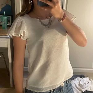 Vit glittrig blus med volangärmar - Säljer en superfin vit blus med glittrigt material som ger en extra touch. Blusen har korta volangärmar som ger en söt och feminin look. Perfekt för både vardag och fest!