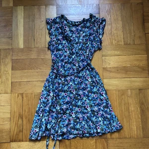 Izabel klänning strl M men mer som xs  - En söt liten sommarklänning med blommigt mönster, knappar och ett litet ”bälte” att knyta runt midjan. Det är små ärmar som sitter mer som ett brett linne. 